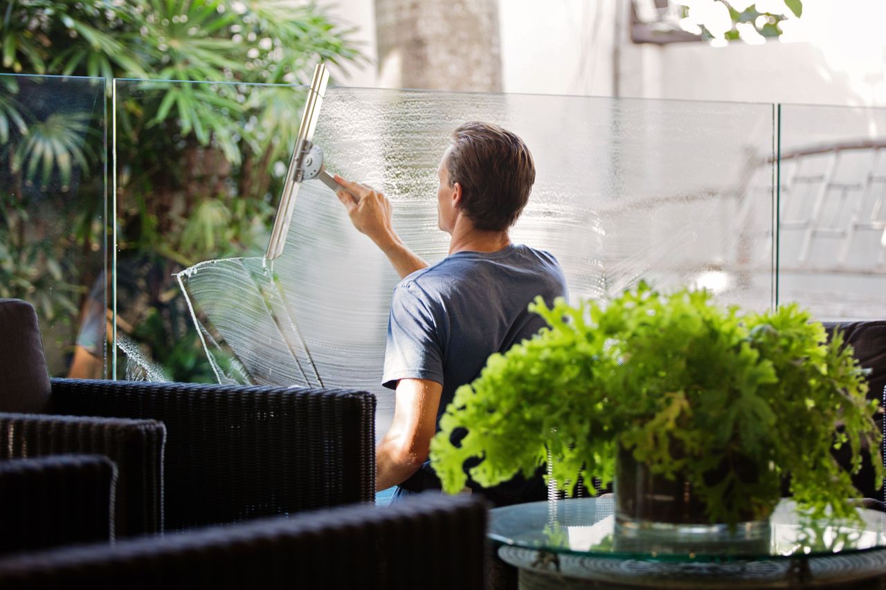team-01 A man cleaning a glass barrier outdoors with a squeegee, surrounded by greenery.